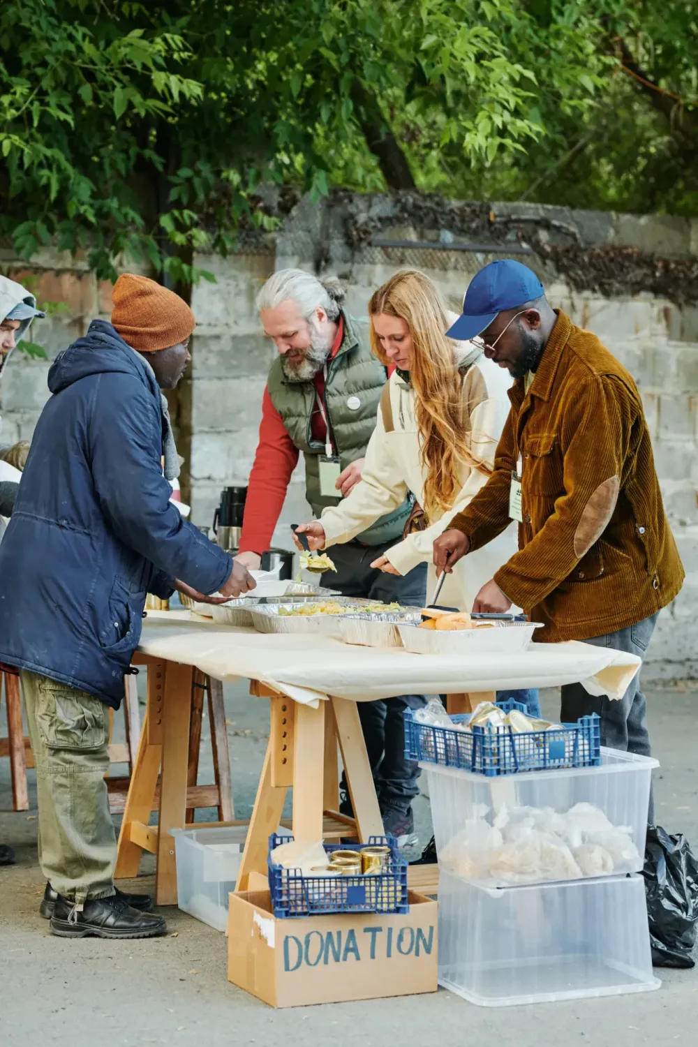 Volunteers serving food from an outdoor table, with a donation box in front